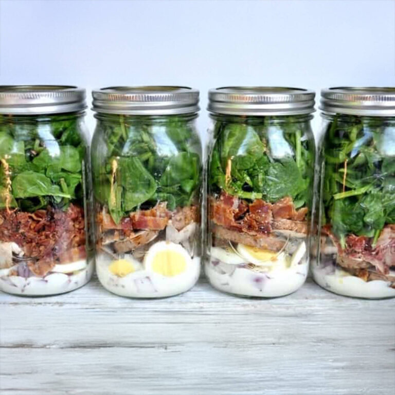 Row of layered salad mason jars with spinach, bacon, and hard-boiled egg against a light blue background