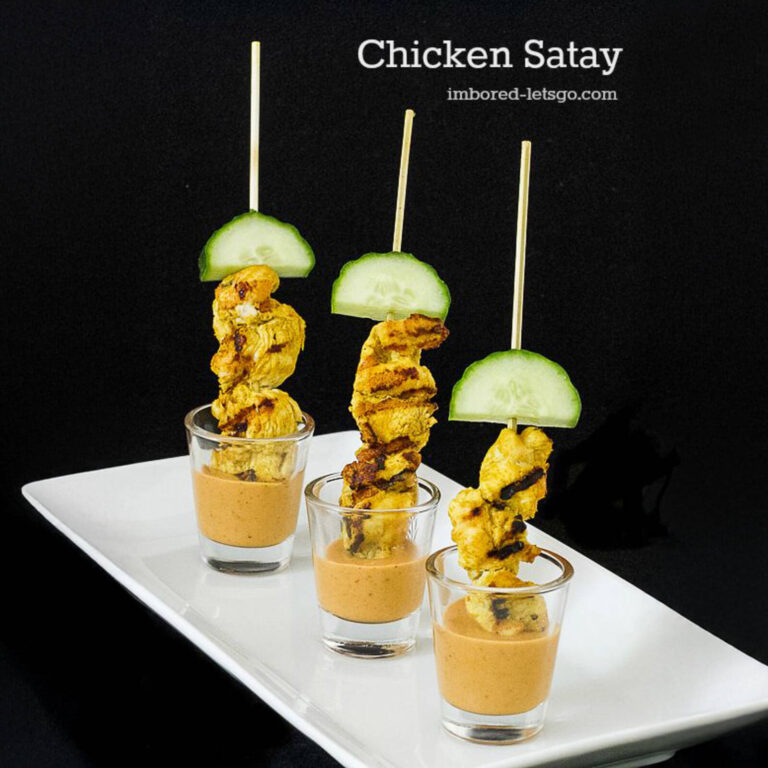 Chicken satay skewers with cucumber slices over peanut sauce shooters, styled on a black background