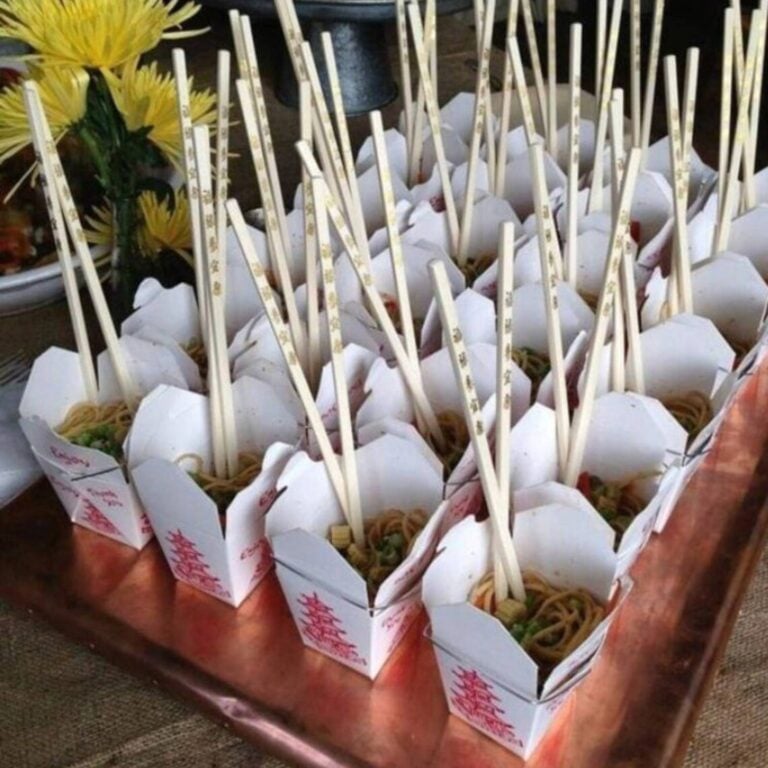 Traditional pagoda-print takeout boxes filled with lo mein noodles, chopsticks, and yellow chrysanthemums on a copper tray