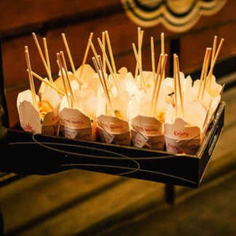 White takeout boxes with handwritten “Enjoy” notes and chopsticks glowing under string lights on a dark tray