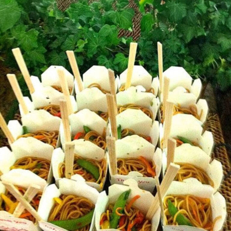 White takeout boxes filled with lo mein noodles and bell pepper, styled outdoors with greenery