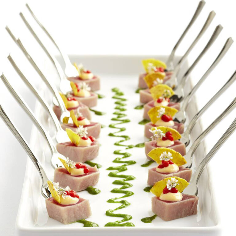 Seared tuna cubes on forks topped with wasabi cream, crispy chip, and pomegranate seeds