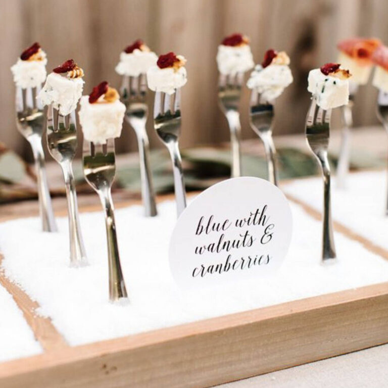 Blue cheese cubes with candied walnuts and cranberries on tasting forks in a snow-themed display labeled “blue with walnuts & cranberries”