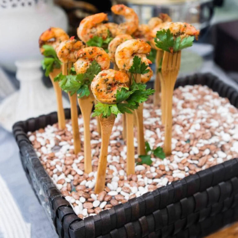 Seasoned grilled shrimp with parsley on wooden tasting forks displayed in a tray of dried beans