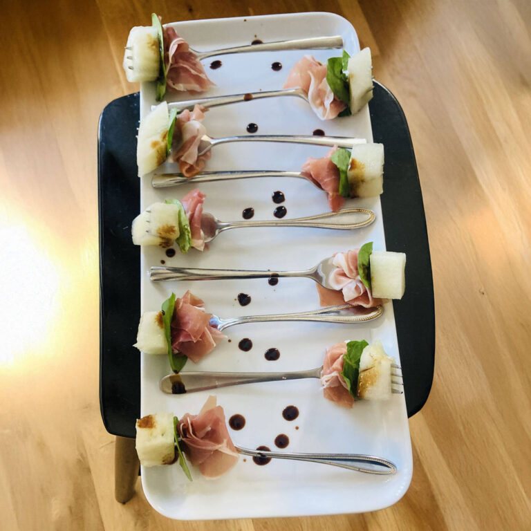 Prosciutto-wrapped melon and basil bites on forks, drizzled with balsamic