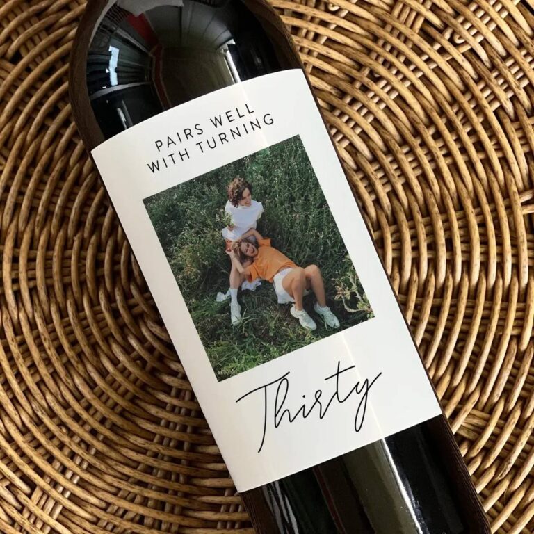 Wine bottle on a woven placemat with a “Pairs Well With Turning Thirty” label featuring two friends laughing together in a grassy field