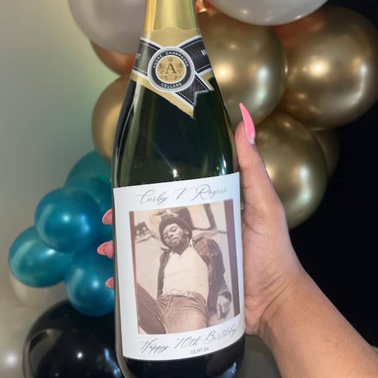 Hand holding a champagne bottle with a “Curby V. Rogers, Happy 70th Birthday, 12.07.24” label featuring a sepia-toned portrait, gold and teal balloons behind