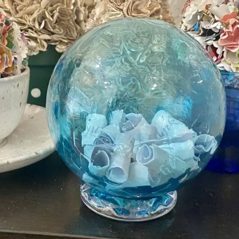 Teal glass wishing ball filled with rolled wish papers, displayed among paper flower arrangements